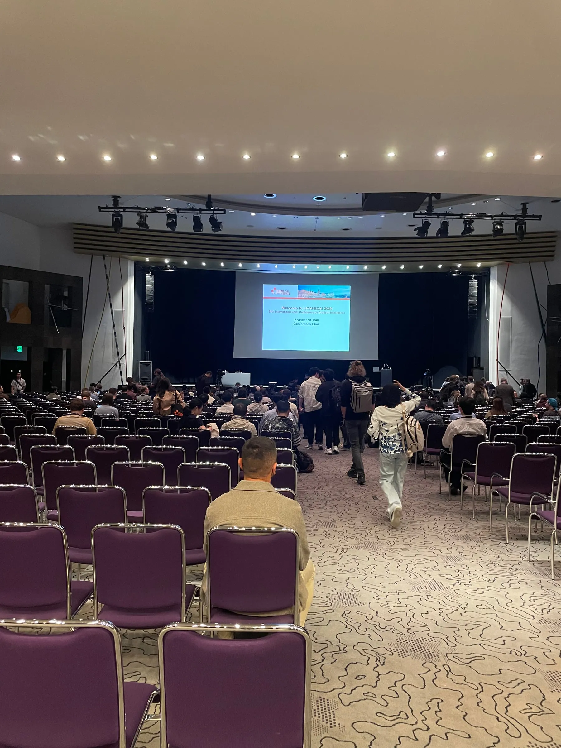 A conference auditorium with purple seats, people taking their places in front of a stage and a large screen